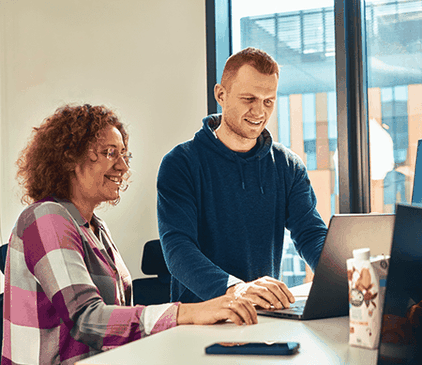 Dwie osoby uśmiechające się podczas pracy na laptopie przy jasnym biurku, z kartonem soku w pobliżu i dużym oknem w tle.