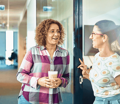 Dwie kobiety uśmiechają się i rozmawiają w korytarzu biurowym. Jedna trzyma kubek z kawą, a druga gestykuluje ręką w pobliżu szklanych drzwi.
