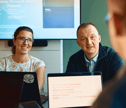 Dwie osoby siedzące przy stole z laptopami, zaangażowane w dyskusję. Prezentacja jest wyświetlana na ekranie w tle.