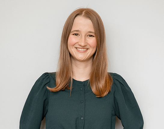 Uśmiechnięta dziewczyna z długimi brązowymi włosami, ubrana w ciemnozieloną bluzkę, stojąca na jasnym tle.