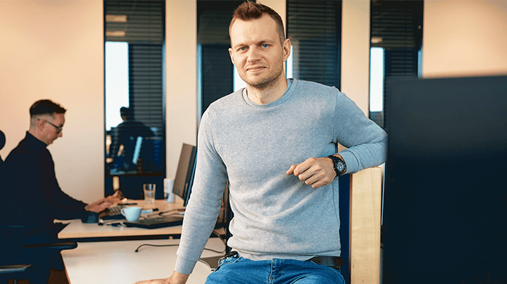 Mężczyzna w szarym swetrze opiera się o biurko w nowoczesnym biurze, z inną osobą pracującą przy komputerze w tle.