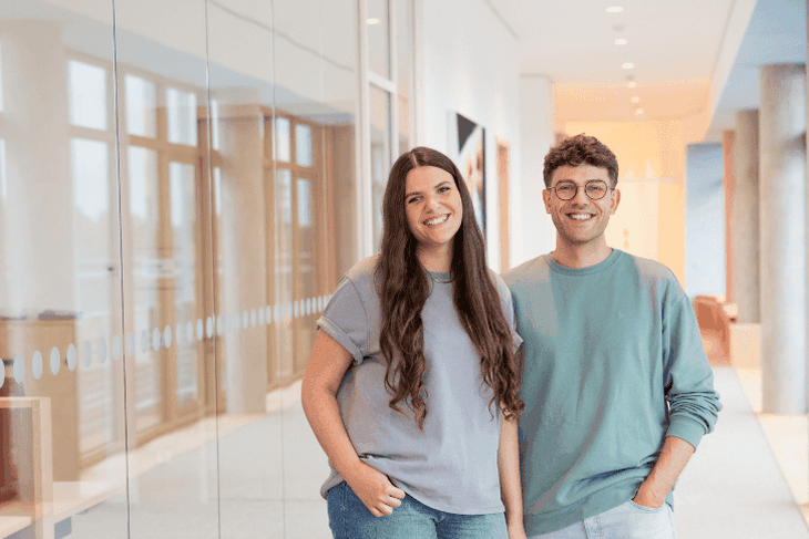 Eine duale Studentin und ein dualer Student stehen im Flur des dm-Unternehmenssitzes in Karlsruhe und lächeln in die Kamera.